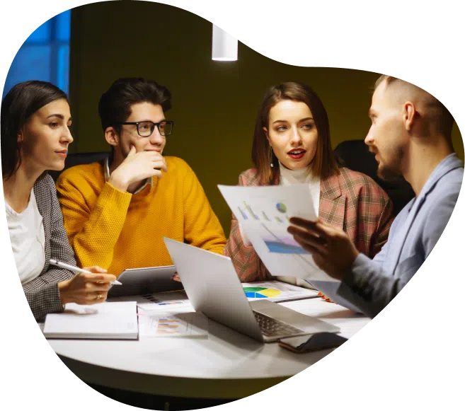 A group of four people in a meeting room are engaged in a discussion. They are seated around a table with open laptops and documents. One person is holding a paper with charts, while the others listen attentively. The room has dim lighting with a single overhead light.