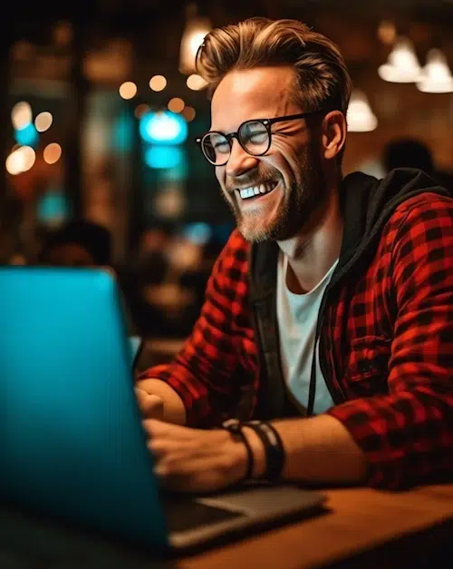 A smiling person with glasses and a beard, wearing a red plaid shirt and a black hoodie, is working on a laptop in a warmly lit café. The background is softly blurred with glowing lights, creating a cozy atmosphere. They appear focused, possibly crafting the next big thing for Envy Websites.
