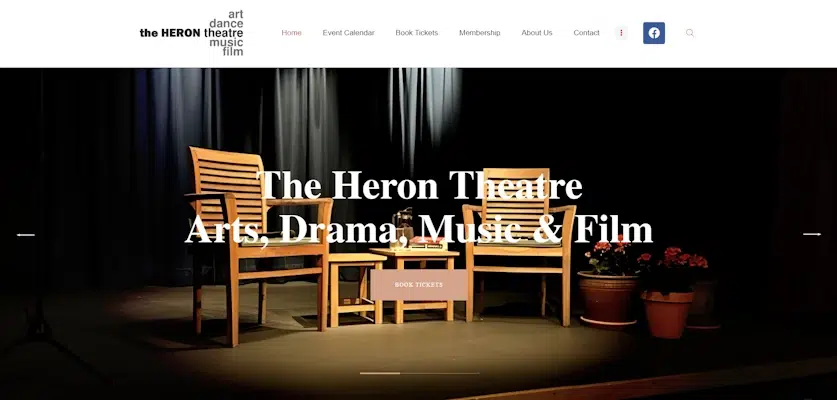 A stage setup at The Heron Theatre with wooden chairs and a table, illuminated by stage lights, surrounded by dark curtains. The text reads "The Heron Theatre Arts, Drama, Music & Film" with a navigation bar featuring Home, Event Calendar, Book Tickets, Membership, About The Heron Theatre, and Contact.